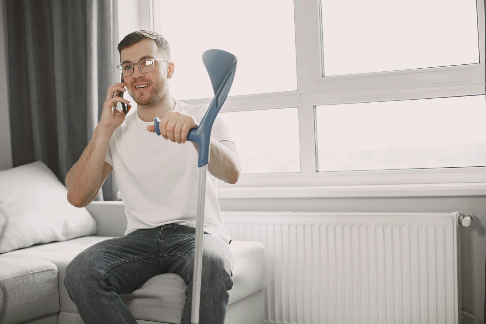 Adult man sitting on couch using crutch, talking on phone in a well-lit room.